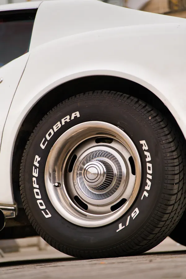 Gros plan sur la roue d'une voiture américaine classique blanche équipée d'un pneu Cooper Cobra Radial G/T à lettres blanches en relief. La jante en acier chromé présente un design vintage avec des ouvertures circulaires et un enjoliveur central brillant.