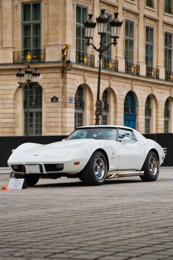 Chevrolet Corvette C3 blanche de collection exposée sur pavés devant façade classique parisienne en pierre de taille avec arcades, balcons dorés et lampadaires d'époque.