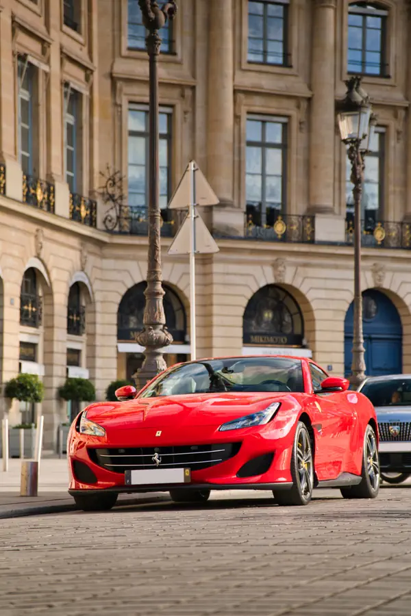 Ferrari Portofino rouge garée devant façade néoclassique parisienne en pierre de taille avec colonnes, arcades et balcons ornés de ferronneries dorées, place Vendôme.