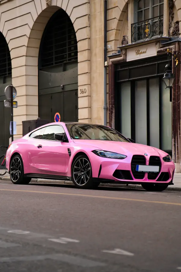 BMW M4 rose vif garée devant un immeuble parisien en pierre blonde avec arcades et balcon en fer forgé, jantes noires contrastantes et calandre BMW caractéristique.