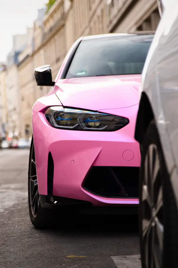 Gros plan sur l'avant d'une BMW M4 rose avec phares LED à accents bleus, pare-chocs sportif et jantes noires, photographiée dans une rue pavée avec architecture haussmannienne en arrière-plan flou.
