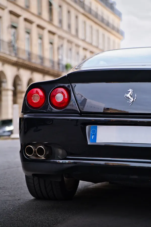 Vue arrière d'une Ferrari noire avec feux ronds rouges caractéristiques, logo cheval cabré et double échappement, devant un immeuble haussmannien.