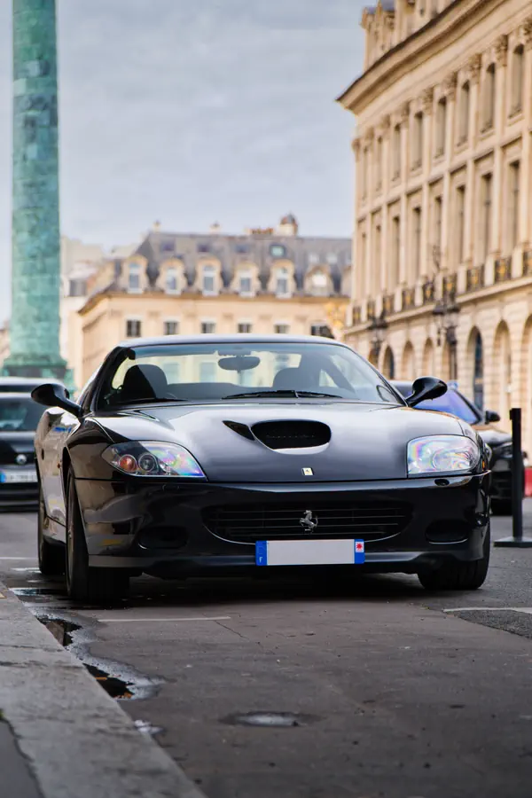 Ferrari 550 Maranello noire vue de face, stationnée devant la place Vendôme avec la colonne et l'architecture classique parisienne en arrière-plan