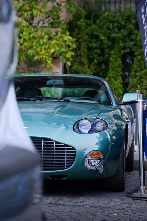 Aston Martin DB7 Zagato verte vue de trois-quarts avant avec phares ronds doubles caractéristiques et calandre horizontale, stationnée dans une cour pavée avec végétation en arrière-plan.