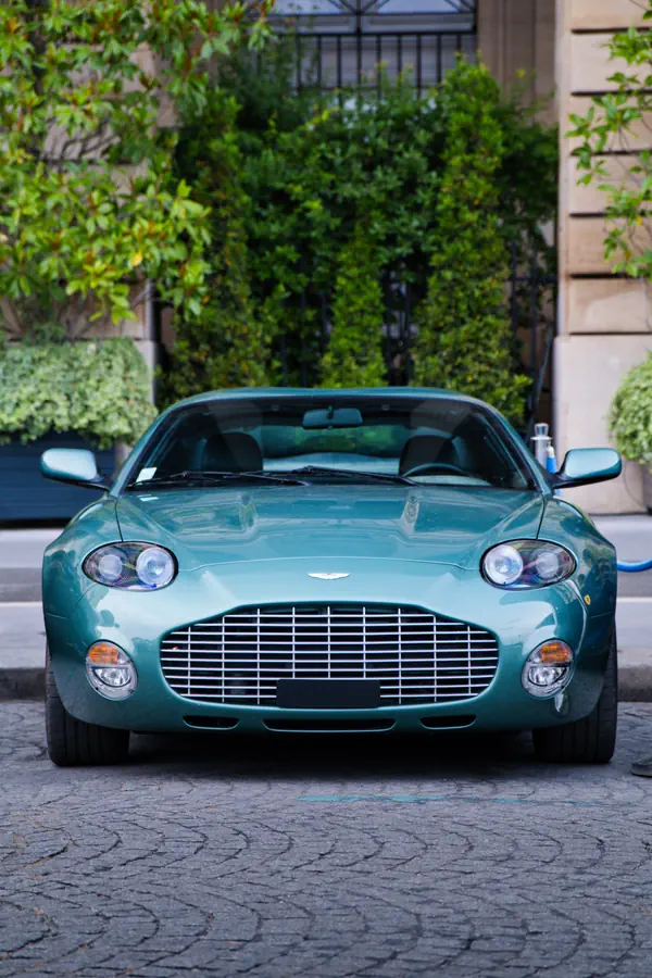 Aston Martin DB7 Zagato verte vue de face avec ses doubles phares ronds et sa calandre horizontale caractéristique, stationnée sur des pavés avec une végétation luxuriante en arrière-plan.