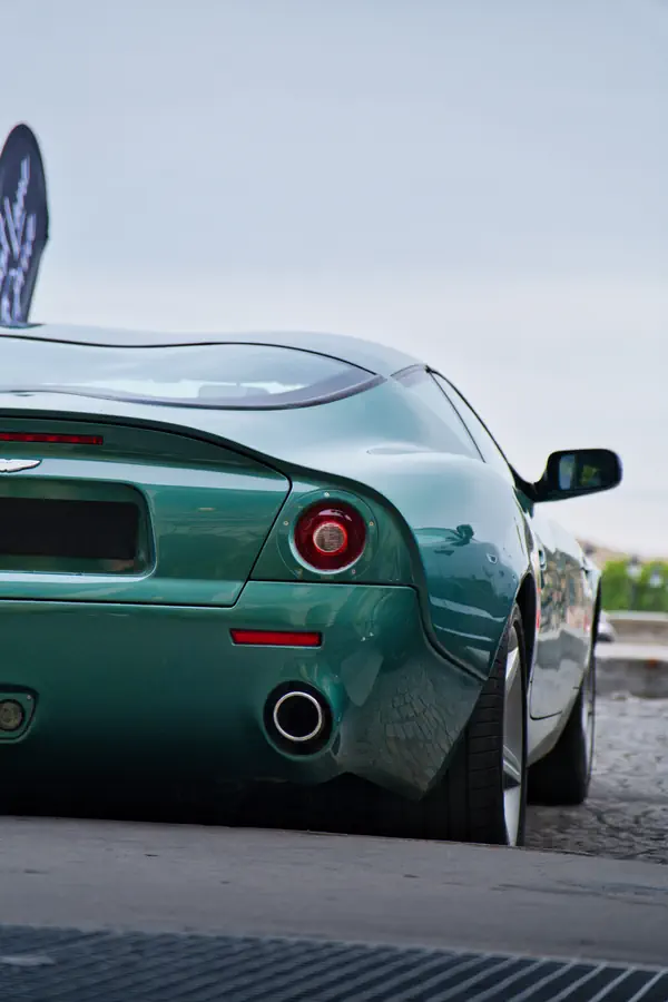 Aston Martin DB7 Zagato verte vue de trois-quarts arrière avec son feu rond rouge caractéristique et son échappement circulaire, sur des pavés avec un ciel nuageux en arrière-plan.