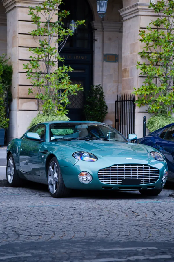 Aston Martin DB7 Zagato verte vue de trois-quarts avant avec ses doubles phares ronds et sa calandre horizontale, stationnée sur des pavés devant un immeuble parisien en pierre avec une végétation soignée.