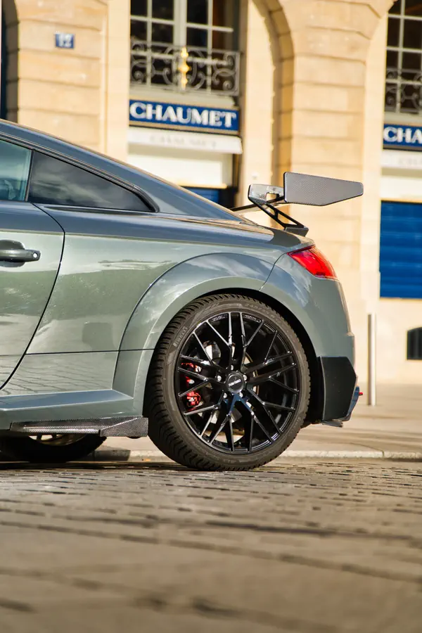 Vue de profil arrière d'une Audi TT RS grise avec aileron en fibre de carbone, jantes noires à rayons et étriers de frein rouges, stationnée devant les boutiques Chaumet.