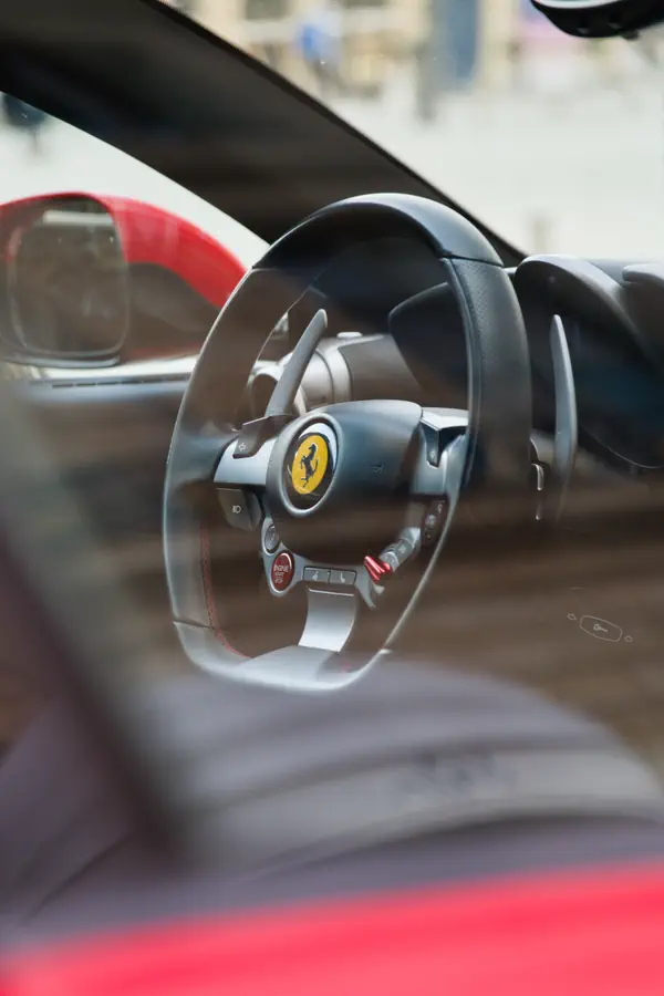 Vue à travers la vitre d'une voiture, gros plan sur le volant en cuir d'une Ferrari avec le logo du cheval cabré au centre et les palettes de changement de vitesse.