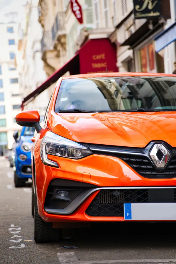Renault Clio orange vif garée dans une rue parisienne, vue rapprochée de l'avant droit, avec un café-tabac et des immeubles en arrière-plan.