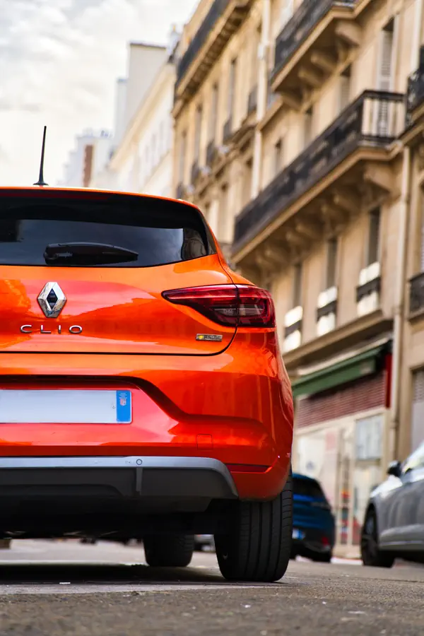 Vue de trois-quarts arrière d'une Renault Clio orange vif garée dans une rue parisienne, avec des immeubles classiques en arrière-plan.