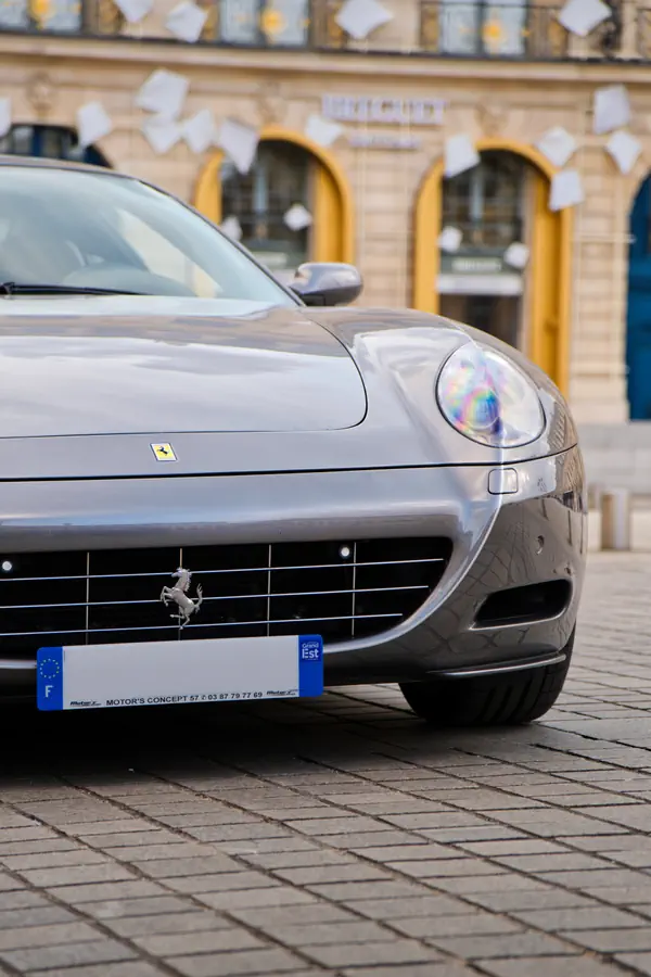 Vue rapprochée de l'avant d'une Ferrari grise garée sur une place pavée, avec en évidence la calandre et l'emblème du cheval cabré, devant un bâtiment à arcades.