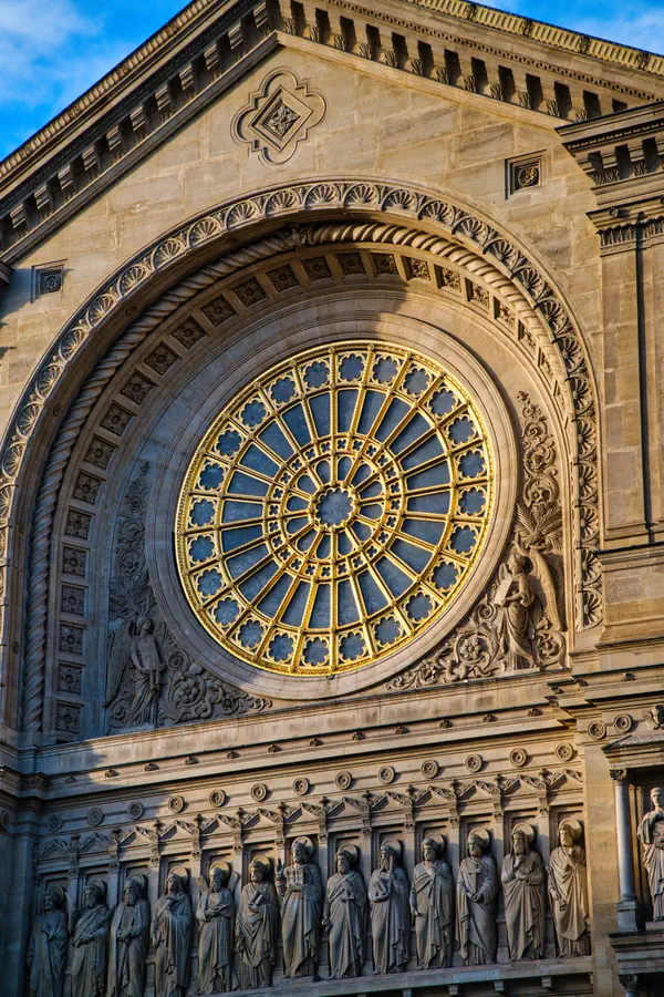 Rosace dorée d'une église de style gothique avec une grande fenêtre circulaire ornementée, encadrée d'arches sculptées et surmontée d'une frise de statues de saints en pierre alignés sur la façade.