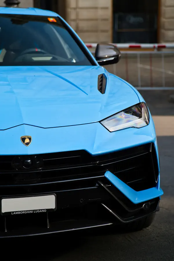 Gros plan sur l'avant gauche d'un Lamborghini Urus bleu ciel, mettant en évidence le logo sur le capot, le phare et le design anguleux du pare-chocs noir.