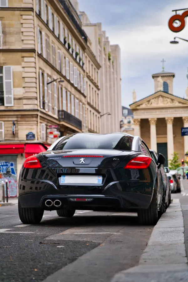 Peugeot RCZ noire garée vue de l'arrière dans une rue parisienne, mettant en évidence son toit vitré à "double bosse" et des immeubles haussmanniens en arrière-plan.