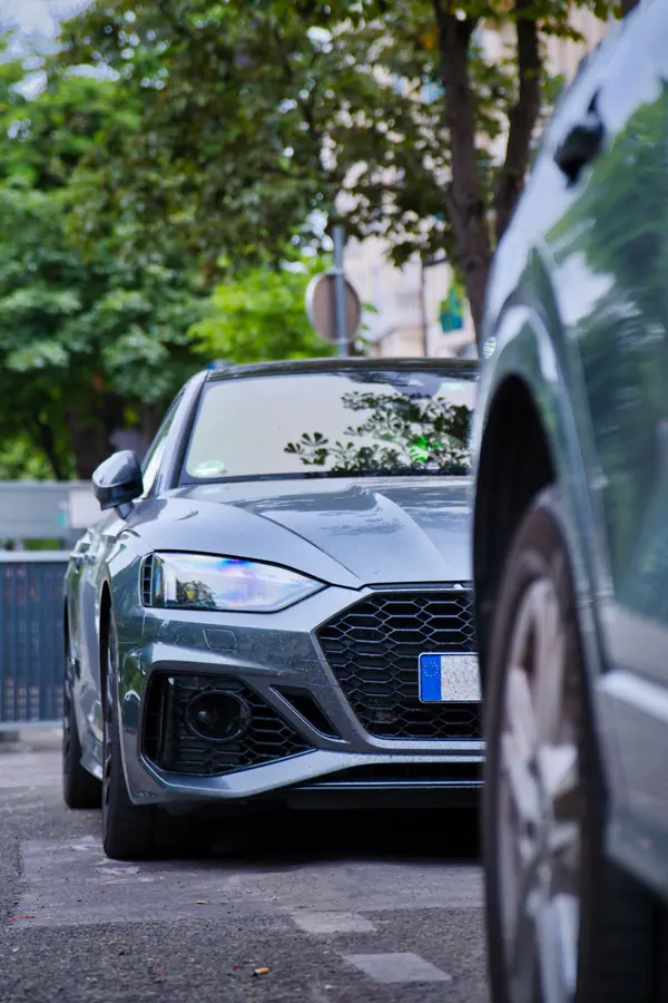 Vue rapprochée de l'avant d'une Audi RS 5 Sportback grise, partiellement cachée par un autre véhicule au premier plan, avec un arrière-plan d'arbres verdoyants.
