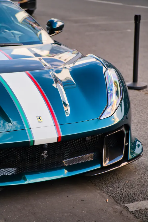 Gros plan sur l'avant d'une Ferrari 812 Competizione de couleur bleu-vert métallisé, avec une bande de course centrale aux couleurs du drapeau italien.