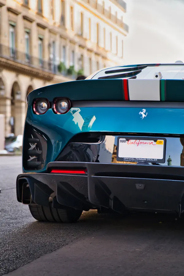 Vue de trois-quarts arrière d'une Ferrari 812 Competizione bleu-vert, mettant en évidence son large aileron avec une bande tricolore et son diffuseur proéminent en fibre de carbone.