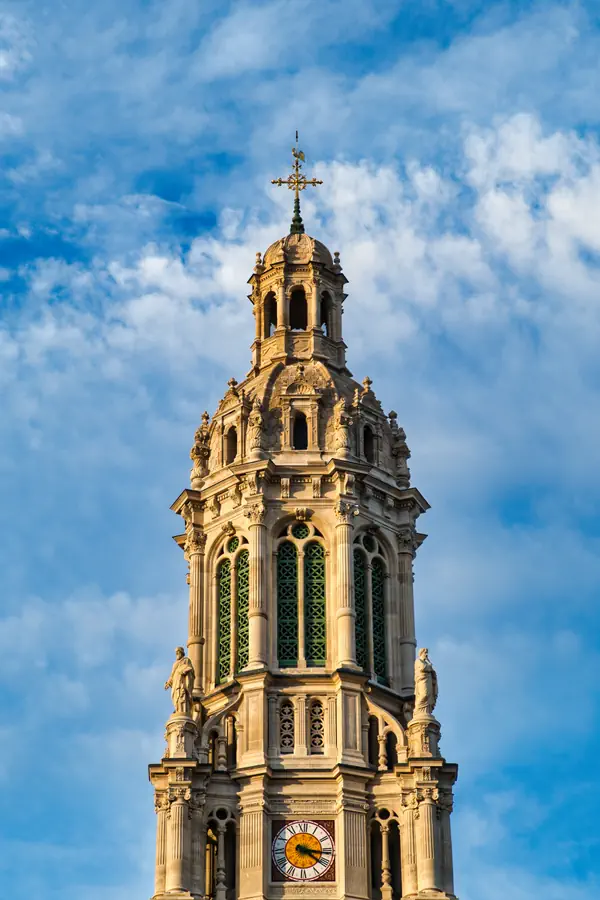 Clocher néogothique en pierre claire orné de vitraux verts, de statues et surmonté d'une croix dorée et d'une girouette. Une horloge à cadran romain est visible sur sa façade, le tout se détachant sur un ciel bleu parsemé de nuages blancs.