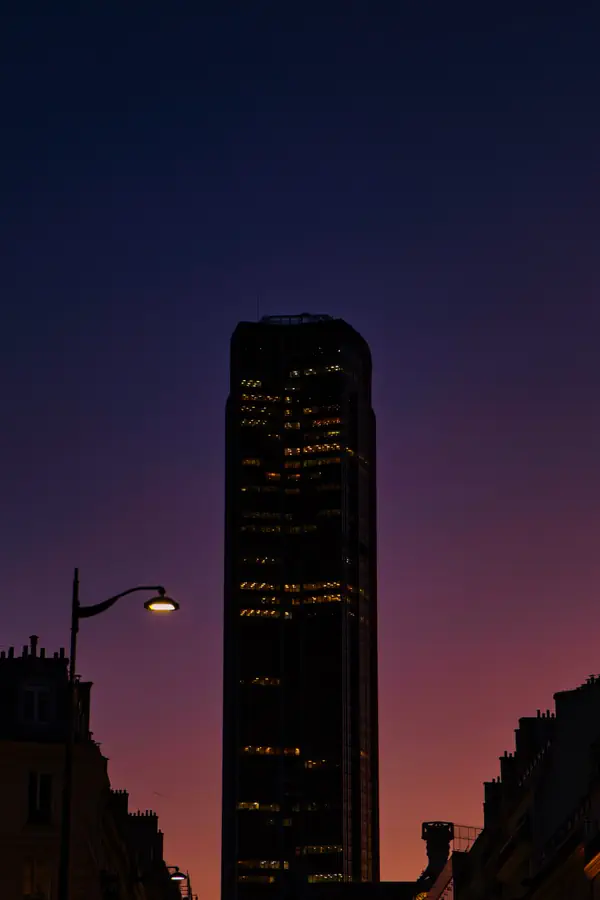 Tour Montparnasse se détachant en silhouette sombre sur un ciel crépusculaire violet et orange, avec quelques fenêtres éclairées et un lampadaire urbain au premier plan.