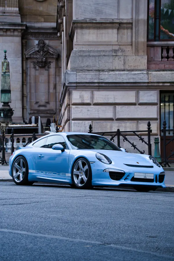 Porsche 911 de couleur bleu clair, avec un kit carrosserie et un lettrage "PORSCHE" sur le côté, garée devant un bâtiment parisien de style classique.