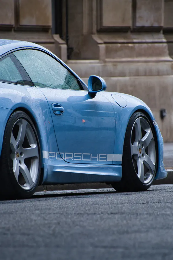Gros plan sur le profil d'une Porsche 911 bleu clair, mettant en évidence le lettrage "PORSCHE" blanc sur le bas de caisse et les jantes argentées à cinq branches.