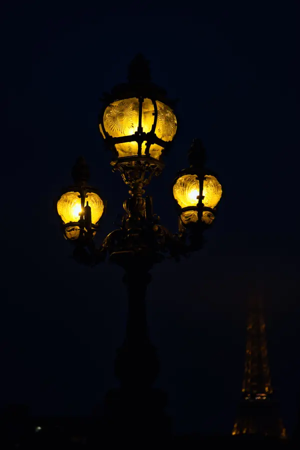 Réverbère parisien ancien à trois lanternes ornementées diffusant une lumière dorée dans la nuit, avec la Tour Eiffel illuminée visible en arrière-plan flou.