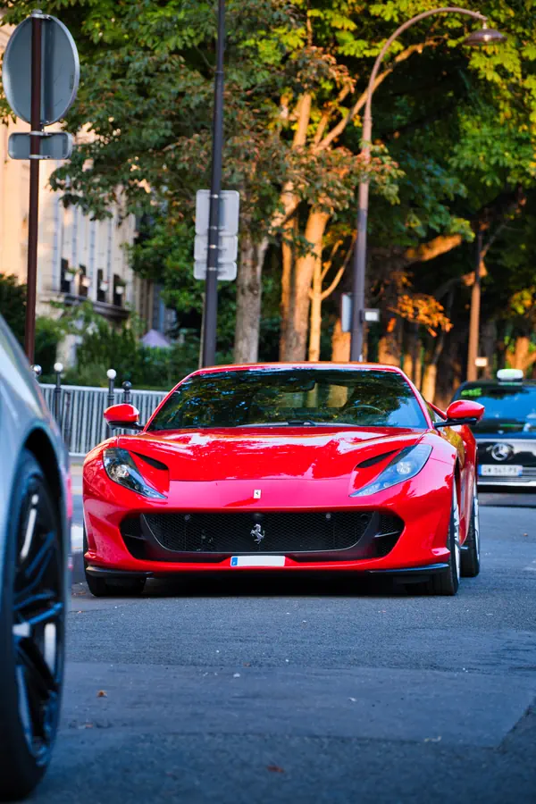 Ferrari 812 Superfast de couleur rouge vif, photographiée de face sur une avenue parisienne bordée d'arbres, avec une autre voiture au premier plan qui cadre l'image.
