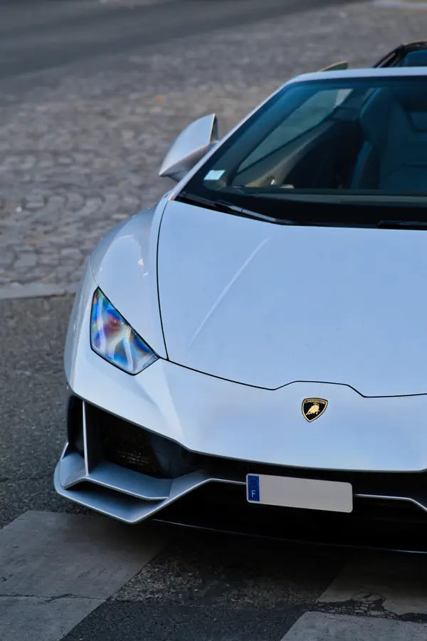 Vue en plongée sur l'avant d'une Lamborghini Huracán Evo Spyder de couleur blanc perle, mettant en évidence le phare, le logo sur le capot et la surface pavée de la rue.