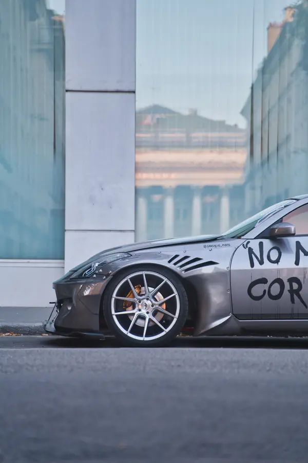 Gros plan sur l'avant d'une Nissan 350Z gris foncé, lourdement modifiée avec un kit carrosserie large, des ailes aérées et des inscriptions de style graffiti sur la portière.