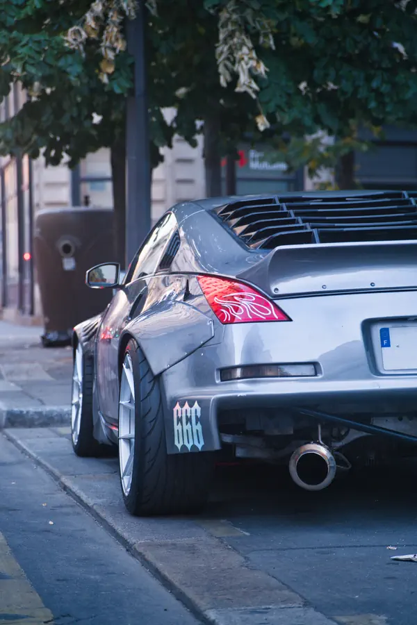 Vue de trois-quarts arrière d'une Nissan 350Z grise modifiée, mettant en évidence son aile arrière élargie et rivetée, une large roue, un grand pot d'échappement et des persiennes sur la lunette arrière.