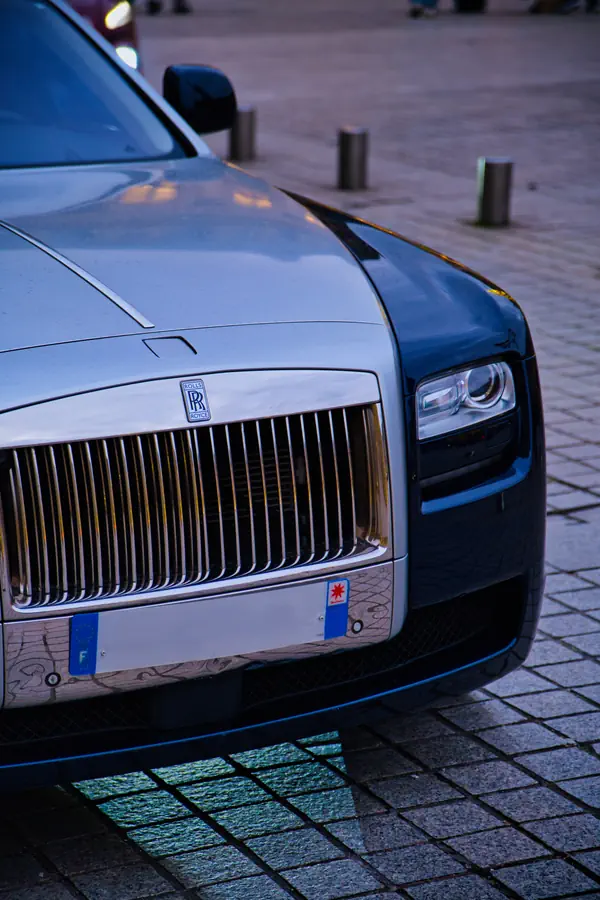 Détail avant d'une Rolls-Royce grise montrant sa calandre chromée caractéristique avec les lamelles verticales, le logo "RR" emblématique, et le phare LED moderne, stationnée sur des pavés urbains.
