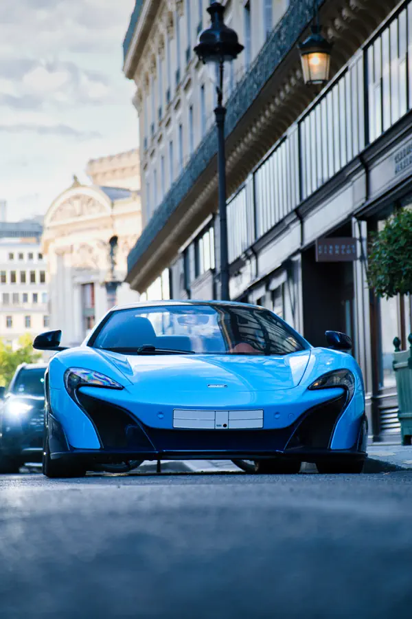 McLaren 675LT de couleur bleu vif, photographiée de face depuis un angle bas dans une rue parisienne, avec des immeubles de style haussmannien en arrière-plan.