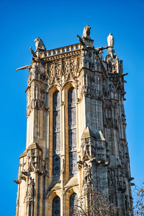 Tour gothique en pierre claire ornée de vitraux, de gargouilles et de sculptures décoratives, avec des pinacles et des statues au sommet, photographiée contre un ciel bleu vif.