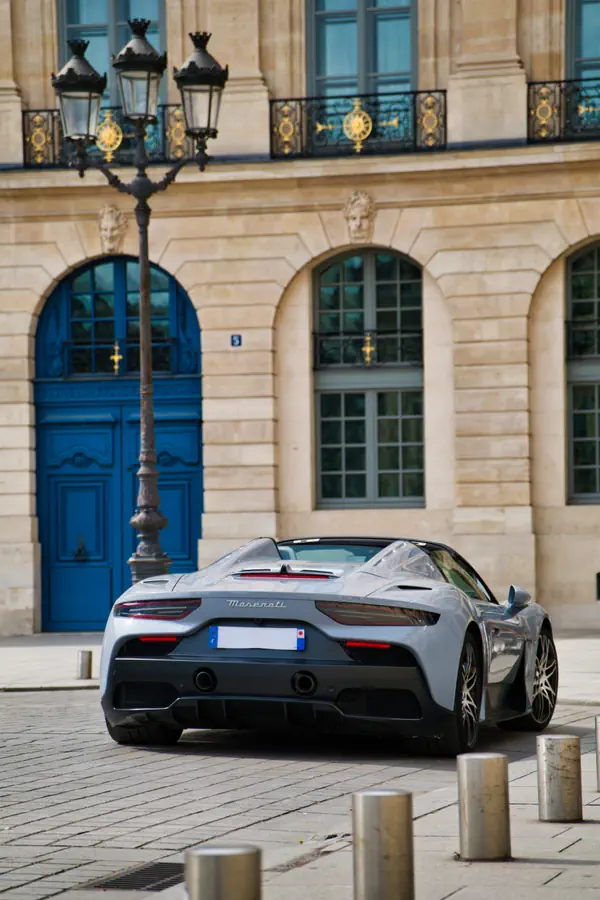 Maserati MC20 Cielo de couleur gris clair, vue de trois-quarts arrière, garée sur une place pavée parisienne devant un immeuble de luxe avec une grande porte bleue.