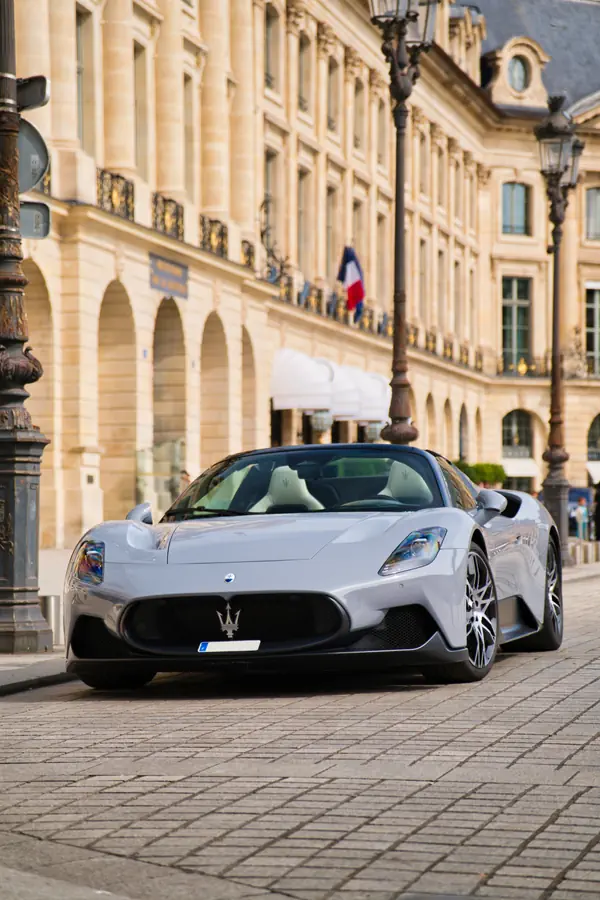 Maserati MC20 Cielo décapotable de couleur gris clair, garée sur les pavés de la Place Vendôme à Paris, avec son intérieur en cuir blanc visible et l'architecture de la place en arrière-plan.