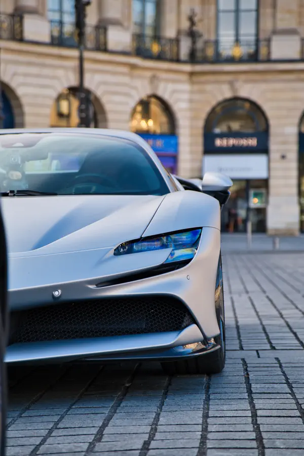 Gros plan sur l'avant d'une Ferrari SF90 Stradale de couleur argent mat, garée sur les pavés de la Place Vendôme, avec un focus sur son phare au design futuriste.