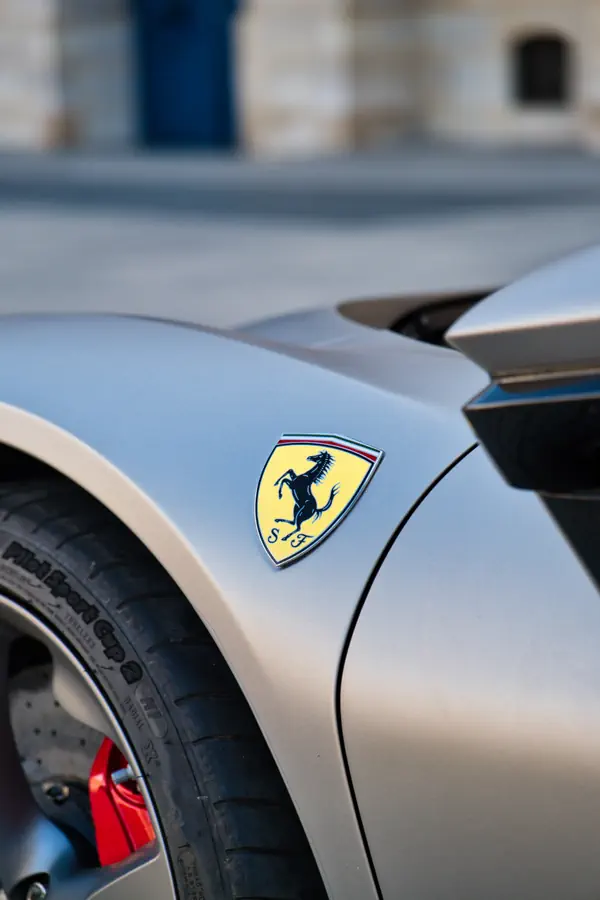 Gros plan sur l'écusson jaune Scuderia Ferrari, le cheval cabré, sur l'aile avant d'une Ferrari SF90 de couleur argent mat, avec une partie de l'étrier de frein rouge visible derrière la roue.