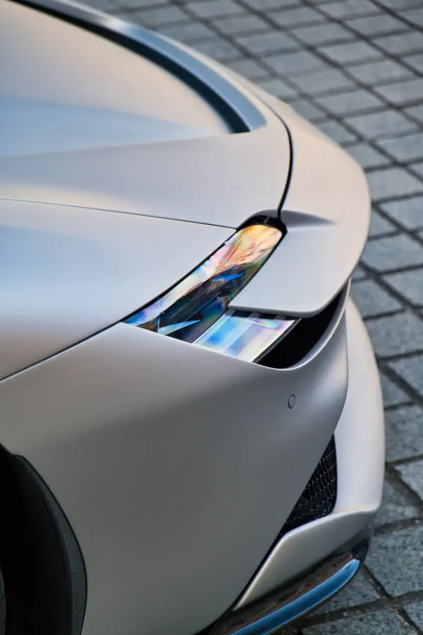 Gros plan sur le phare au design fin et futuriste d'une Ferrari SF90 Stradale, mettant en évidence les reflets colorés dans l'optique et la peinture de couleur argent mat de la carrosserie.