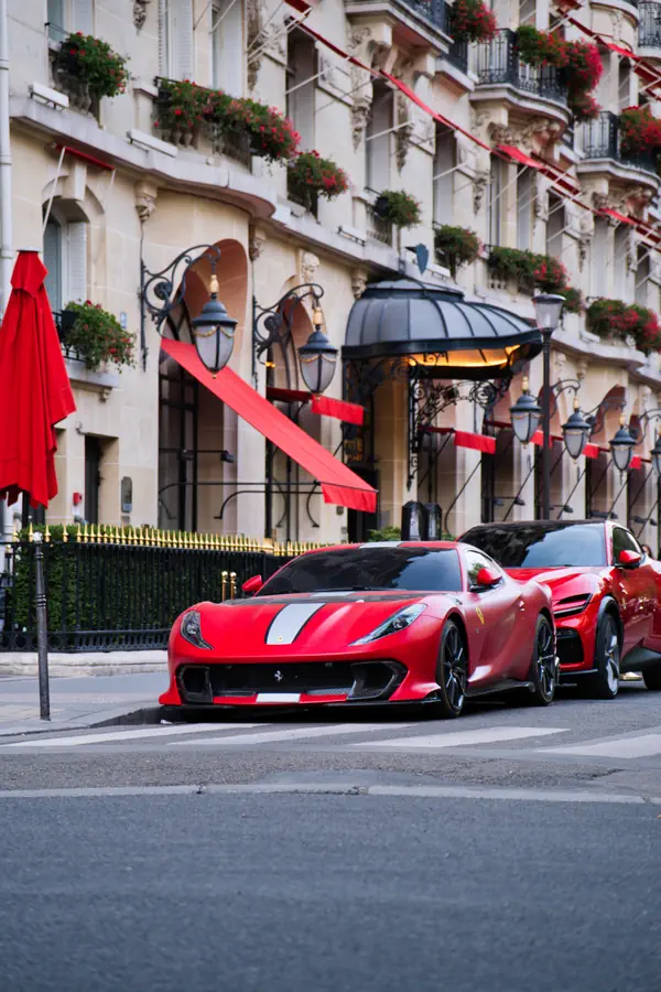 Ferrari 812 Superfast rouge, avec une bande de course centrale, garée devant l'entrée emblématique de l'Hôtel Plaza Athénée à Paris avec ses auvents rouges.