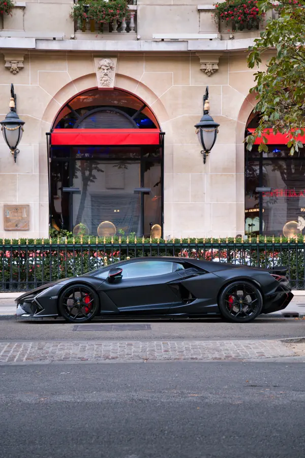 Vue de profil d'une Lamborghini Revuelto de couleur noir mat, avec des étriers de frein rouges, garée dans une rue parisienne devant la façade d'un hôtel de luxe.