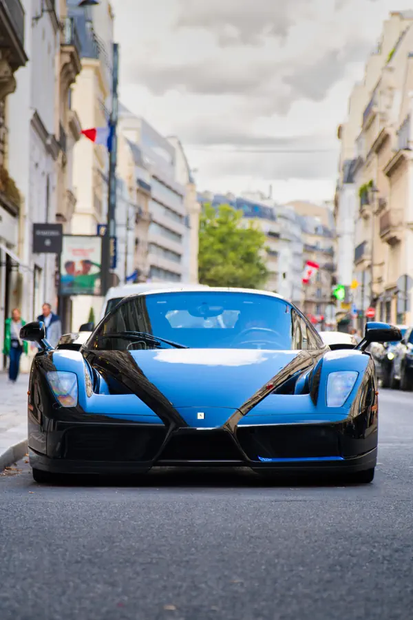Vue de face à ras du sol d'une Ferrari Enzo noire, garée dans une rue parisienne, mettant en valeur son nez pointu inspiré de la Formule 1 et la perspective des immeubles en arrière-plan.