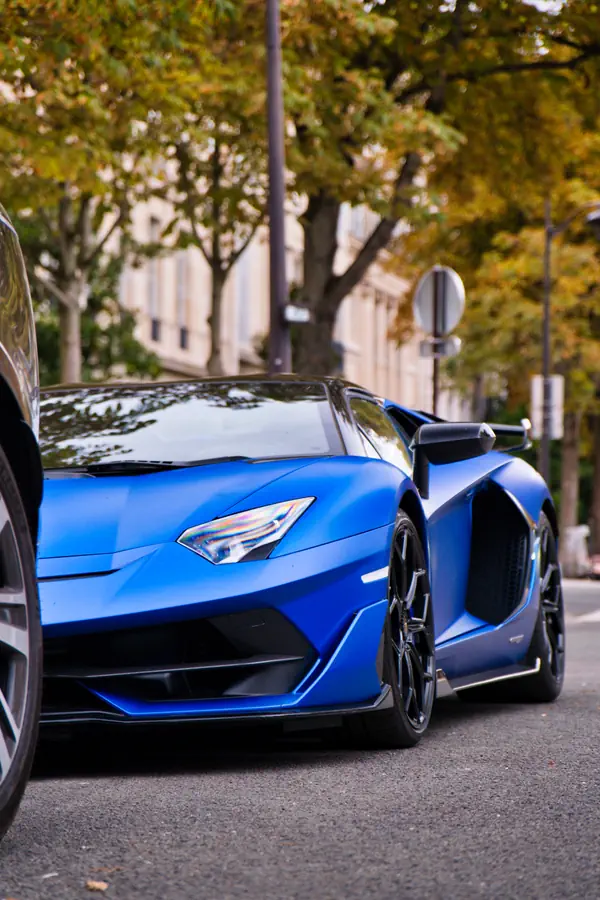 Lamborghini Aventador bleu satiné vue en contre-plongée dans une rue bordée d'arbres et de bâtiments haussmanniens.
