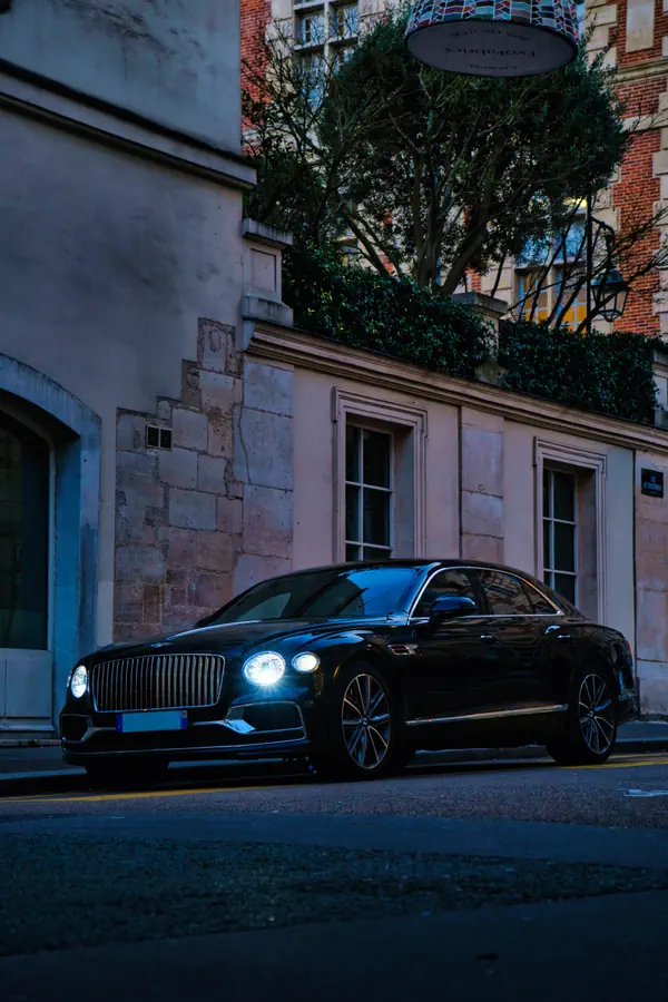 Bentley Flying Spur noire stationnée dans une rue parisienne au crépuscule, ses phares LED allumés illuminant la façade d'un bâtiment en pierre de taille.