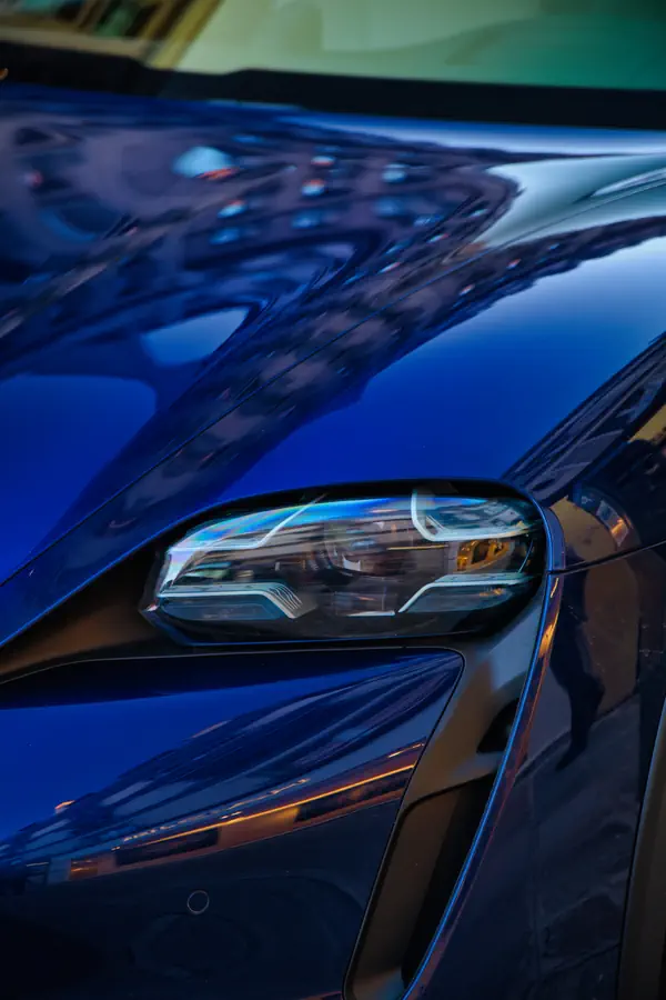 Gros plan sur le phare avant LED d'une Porsche Taycan de couleur bleue foncée, montrant le design sophistiqué et futuriste de l'optique ainsi que les lignes fluides et aérodynamiques de la carrosserie.
