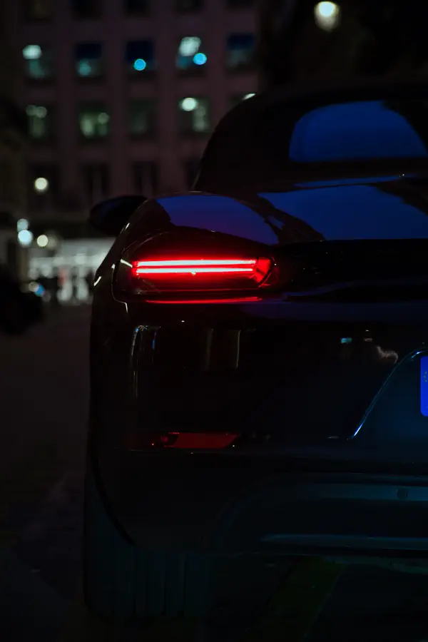 Feu arrière LED d'une voiture de sport dans l'obscurité, montrant une bande lumineuse rouge horizontale distinctive qui se détache sur la carrosserie sombre, avec des reflets bleutés et des lumières de la ville en arrière-plan flou.
