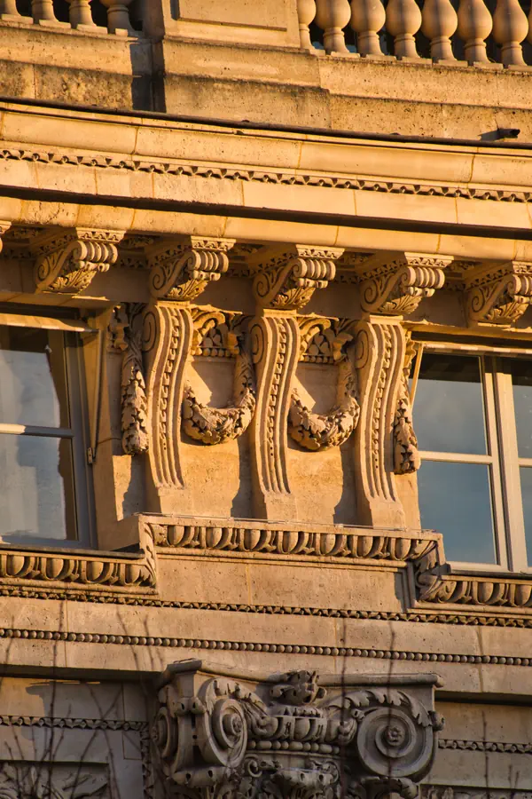 Détails ornementaux d'une façade classique en pierre, mettant en relief des modillons sculptés sous une lumière rasante dorée.
