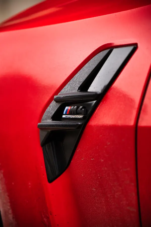 Gros plan sur l'ouïe latérale d'une BMW M4 Competition orange, montrant le badge M Competition intégré dans l'insert noir brillant, avec les couleurs caractéristiques BMW Motorsport (bleu, rouge et bleu marine).
