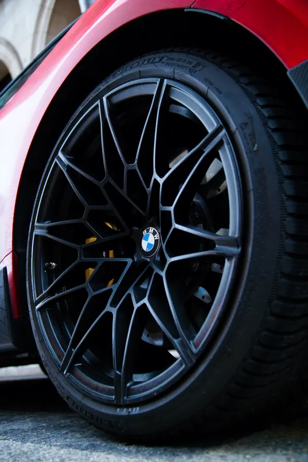 Gros plan sur une jante BMW en alliage noir mat au design complexe en Y, avec le logo BMW au centre et un étrier de frein jaune visible derrière les rayons. La jante est montée sur un pneu Michelin et se détache sur une carrosserie orange.