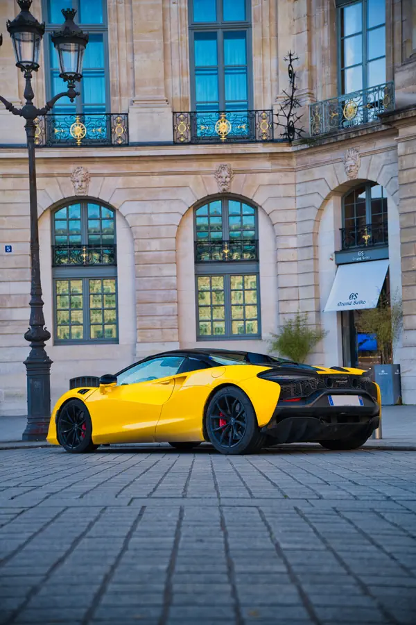 McLaren Artura jaune photographiée de trois-quarts arrière devant une boutique avec des fenêtres cintrées caractéristiques et ses balcons en fer forgé ornés. Le réverbère traditionnel parisien et les pavés au sol complètent cette scène alliant modernité automobile et architecture classique.